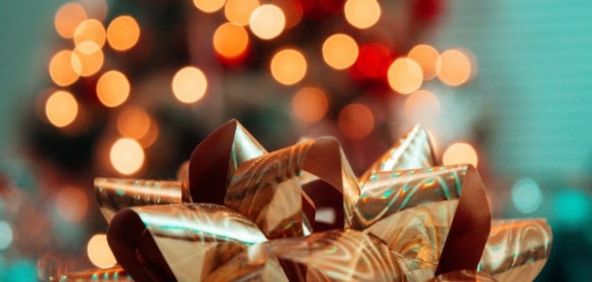 A beautifully wrapped gift bow in the foreground with a blurred Christmas tree adorned with lights in the background.