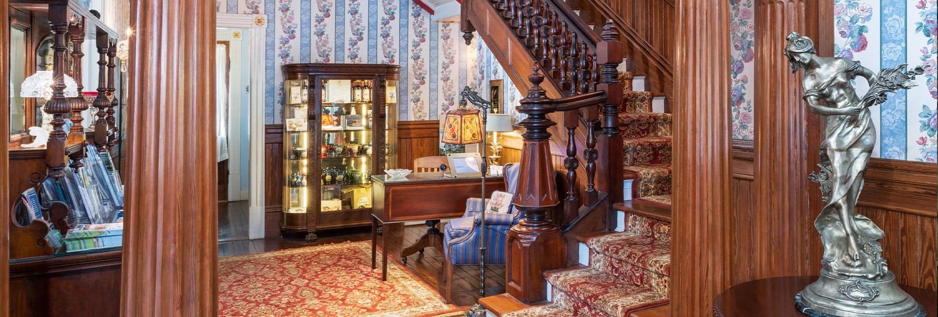 A beautifully decorated entryway featuring ornate wooden staircases, floral wallpaper, and a vintage display cabinet.