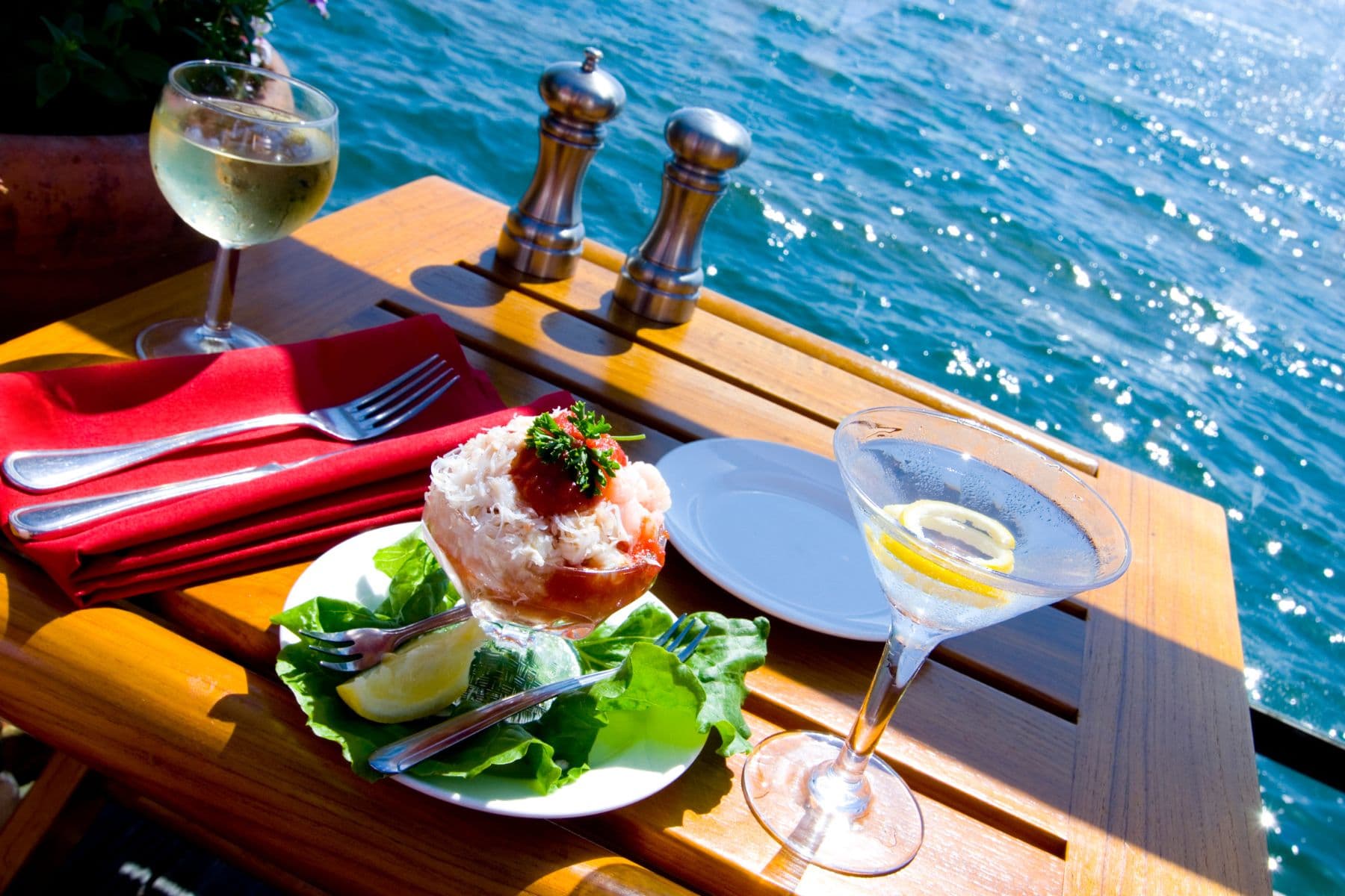A plate of seafood on a wooden table beside a glass of white wine and a martini, with a shimmering water view in the background.