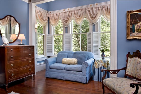 Blue sofa in window box with Floral valances on windows and shutters. Highback chair and antique dresser in foreground.
