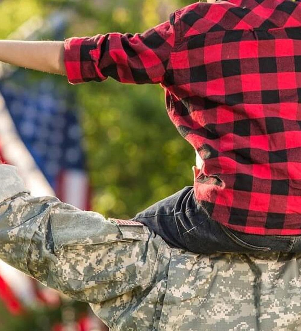 A child in a red plaid shirt rides on a soldier's shoulders with American flags in the background.