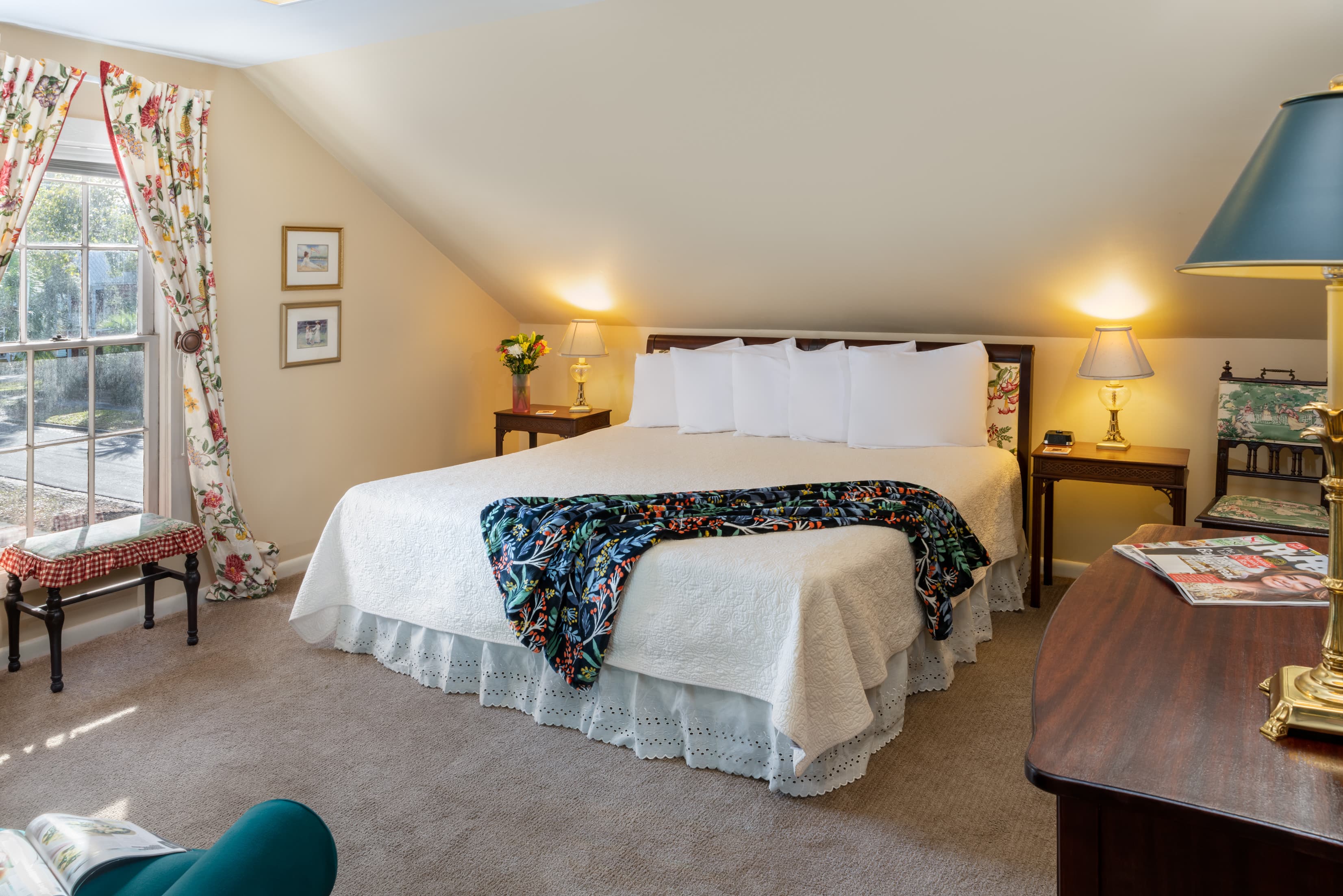 Spacious loft-style bedroom featuring a king bed, soft neutral décor, cozy seating area, floral curtains, and warm natural light