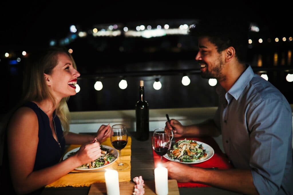A couple shares a joyful dinner outdoors at night, with candles and wine in a romantic setting.