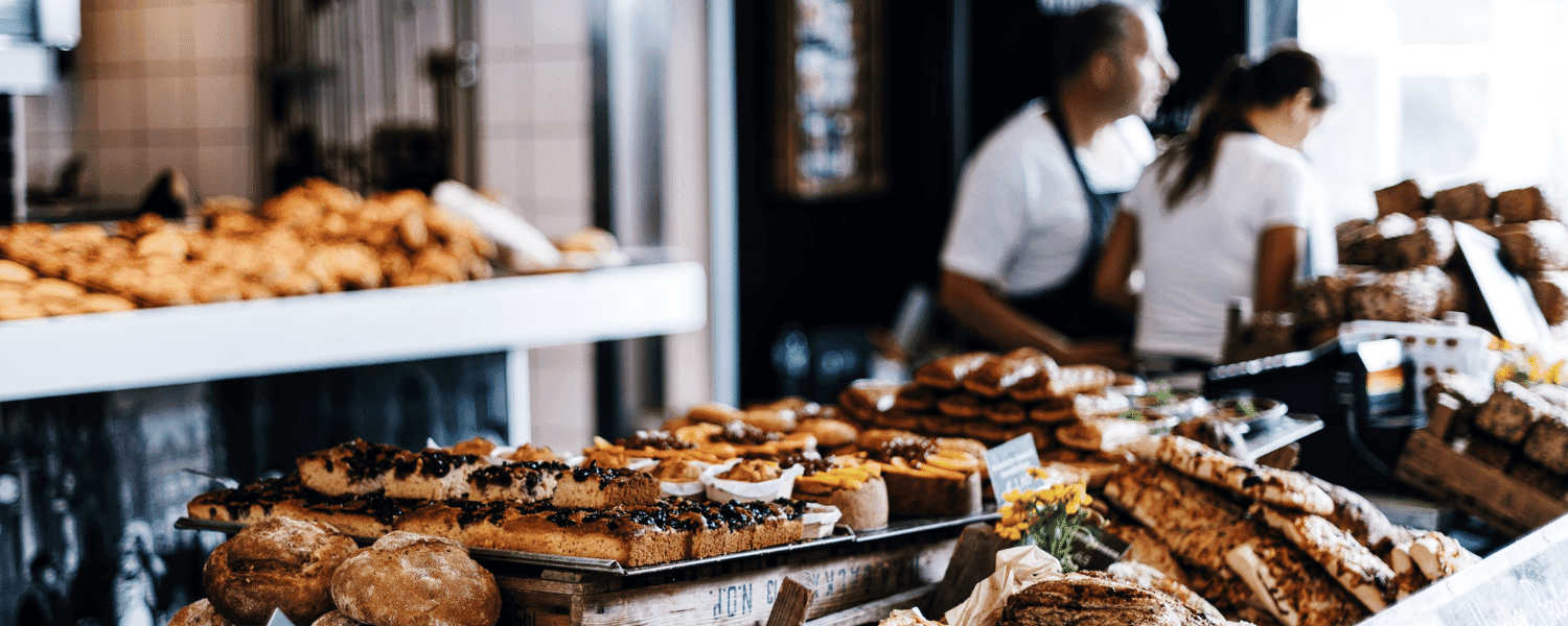 A variety of fresh pastries and baked goods are displayed in a bakery with two employees in the background.