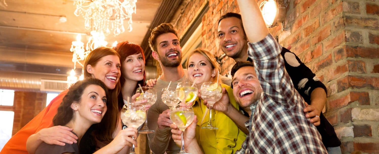 A group of eight friends joyfully raising cocktails while smiling and posing for a photo in a stylish setting.
