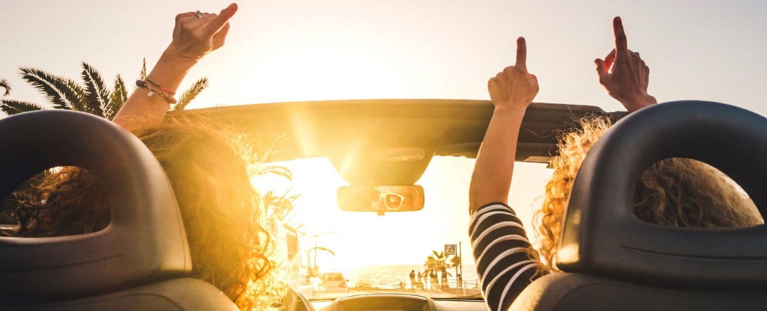 Two people in a convertible raise their arms while enjoying a sunset drive.