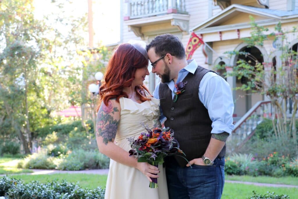 A couple shares a tender moment outdoors, surrounded by greenery, with the woman holding a bouquet.