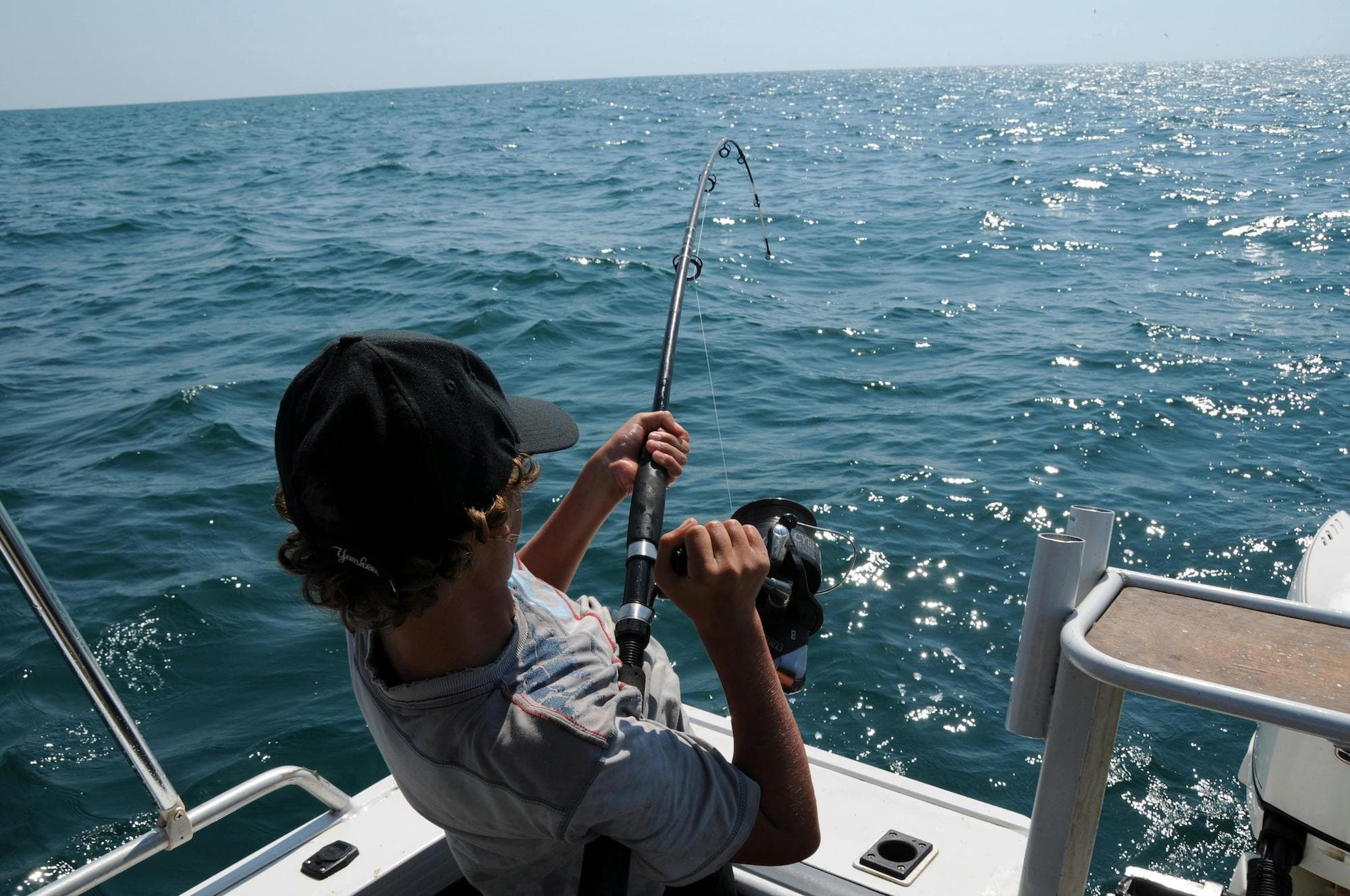 A young person with curly hair is fishing from a boat, reeling in a line over calm blue waters.