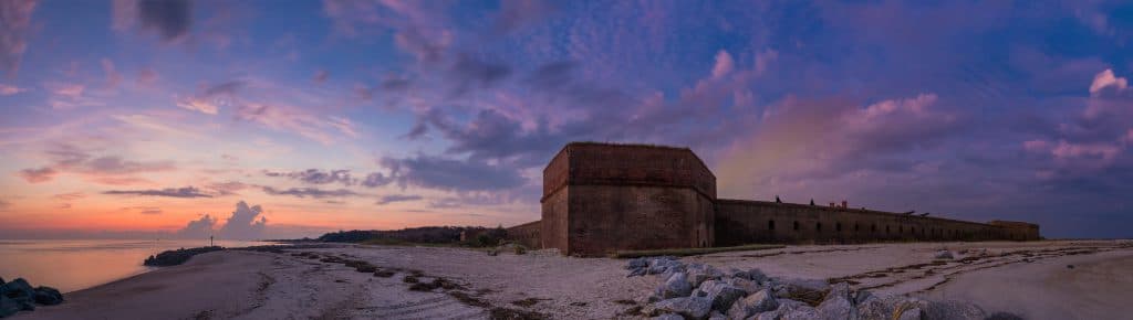 A historic fort overlooking a sandy beach at sunset under a colorful sky.