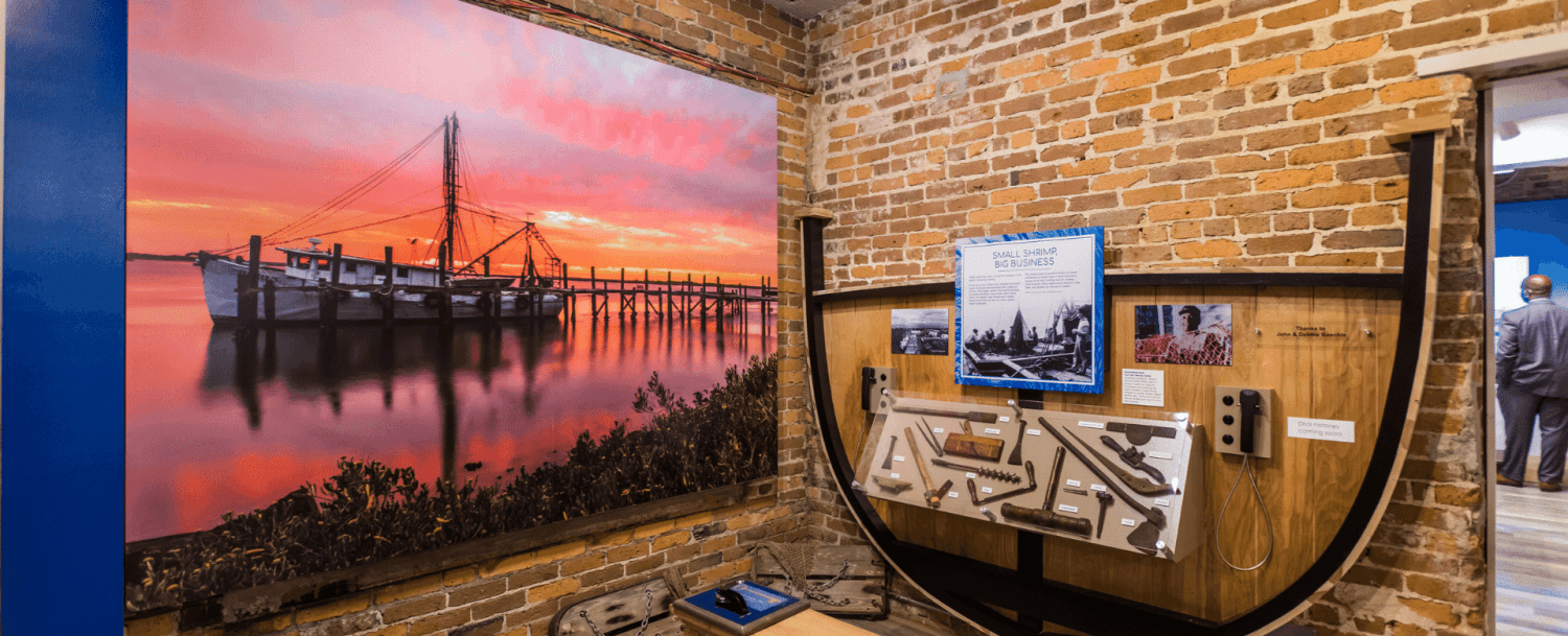 Interior view of a museum exhibit featuring a sunset photograph of a fishing boat and interactive displays about shrimping.