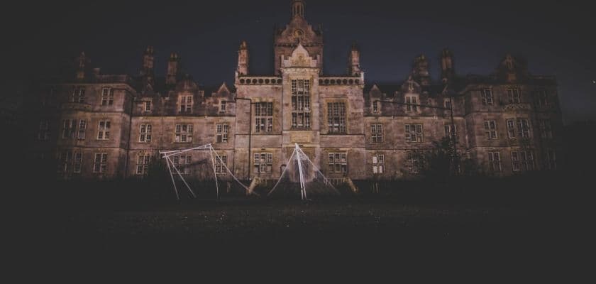 An abandoned mansion looms ominously in the night, with a dilapidated swing set in the foreground.