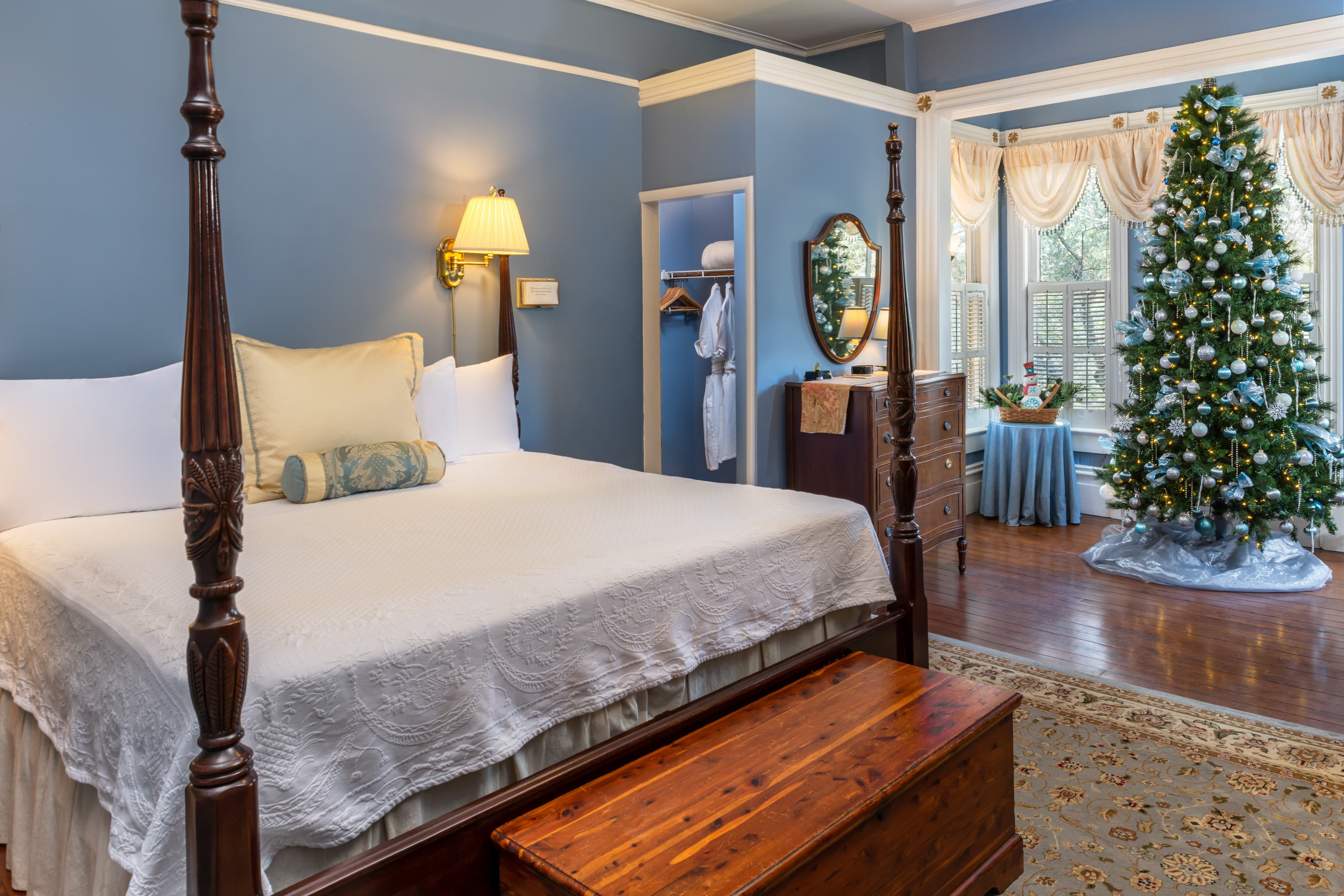 Beautifully decorated holiday guest suite showcasing a four-poster bed, sparkling Christmas tree, bay windows with natural light, hardwood floors, and charming historic furnishings.