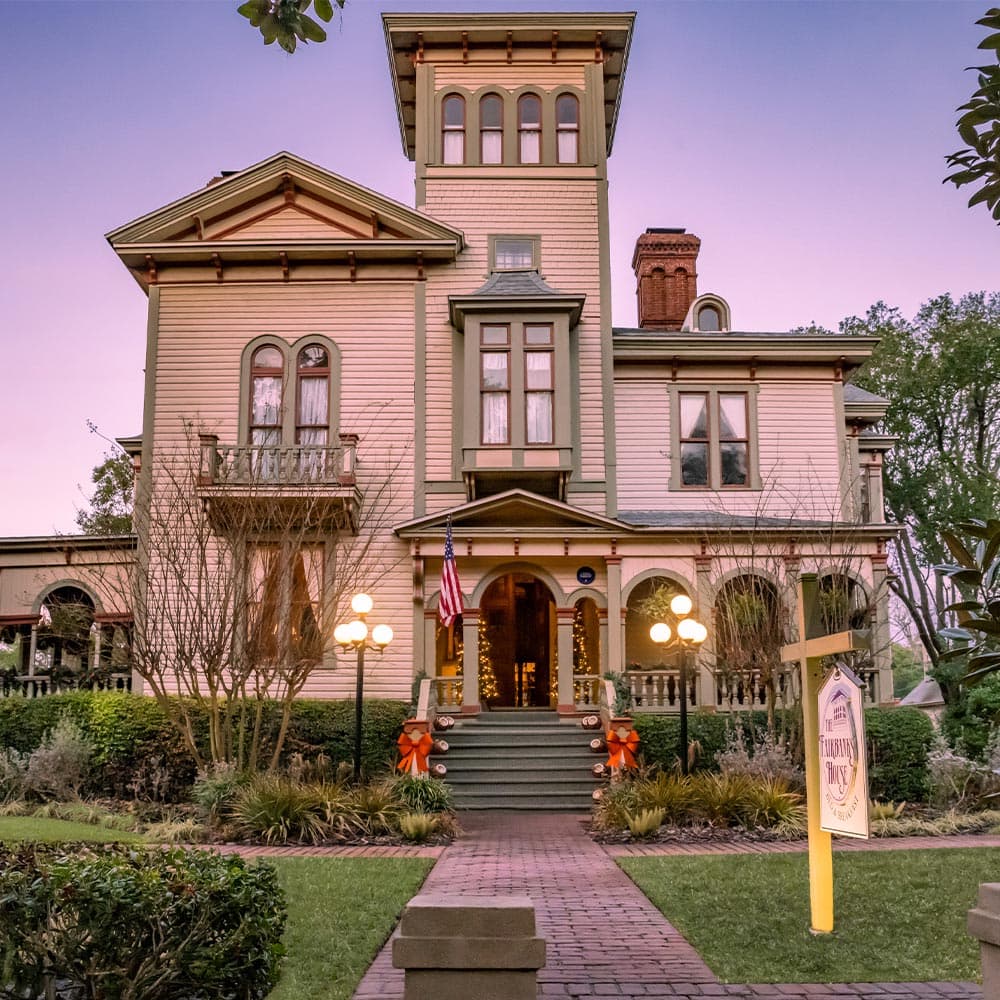 A historic, two-story Victorian-style house with a tower, surrounded by greenery and decorated for the holidays.
