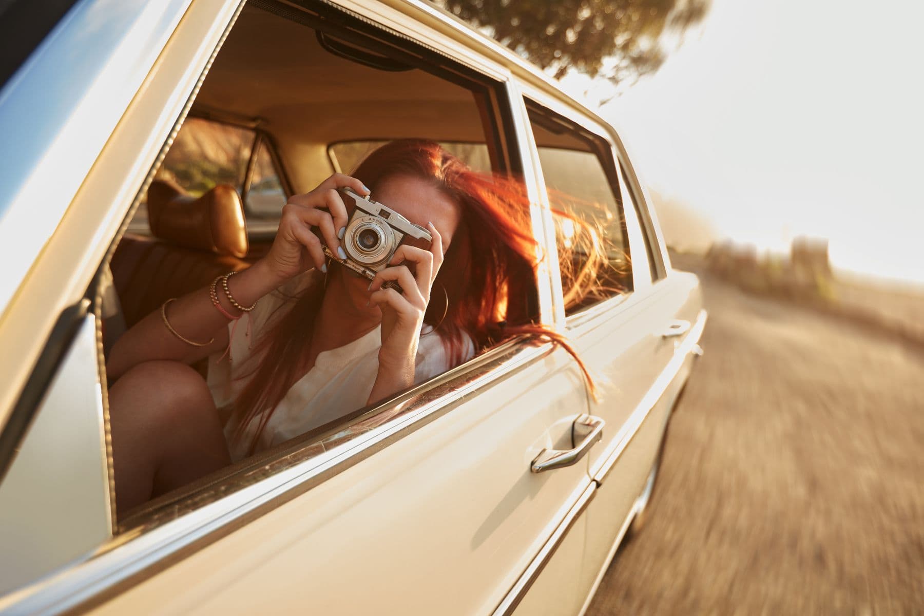 A person with long red hair peers out of a car window, holding a camera.