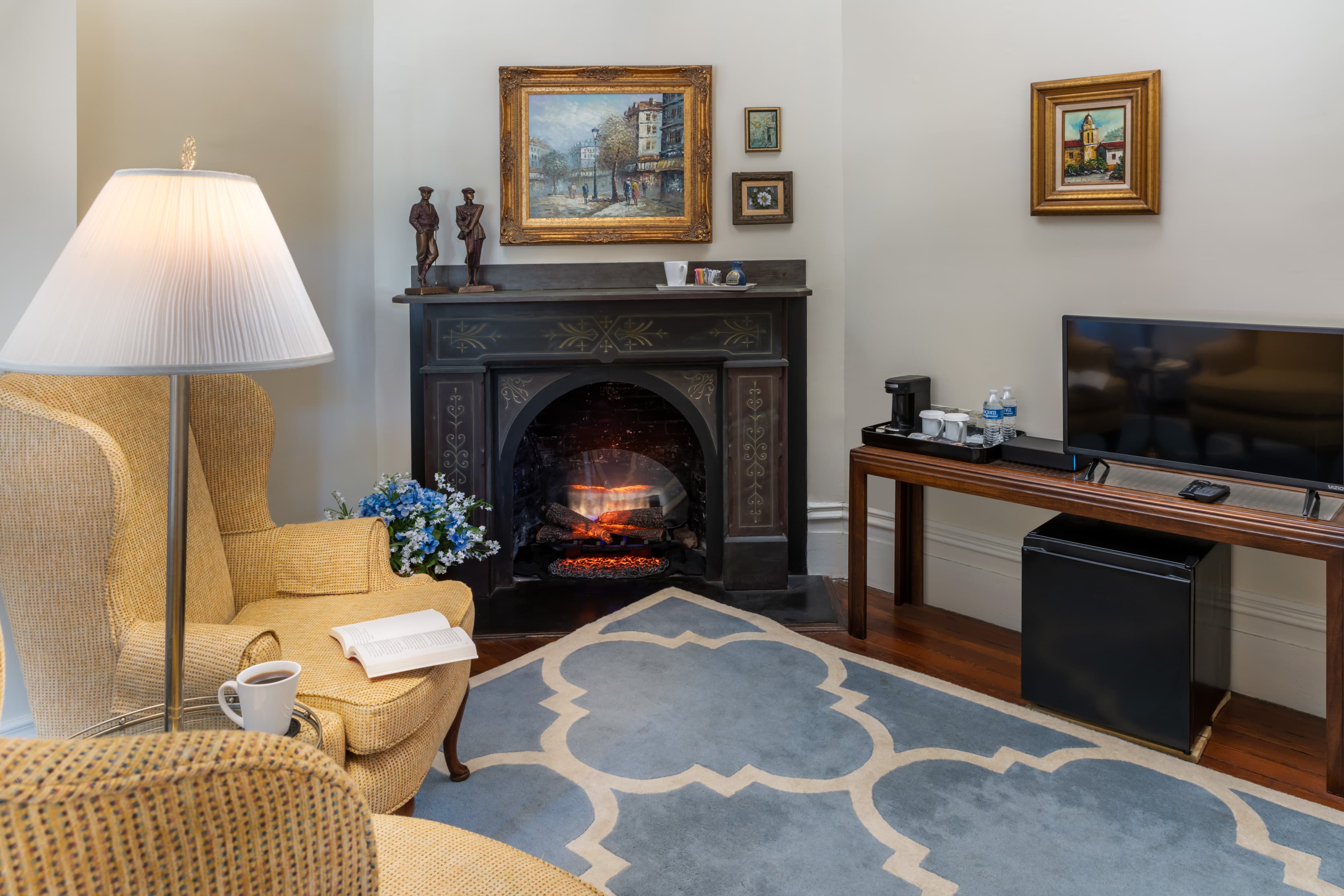 Cozy sitting area inside a guest suite featuring a decorative fireplace, plush armchairs, hardwood floors, vintage artwork, a flat-screen TV, and warm ambient lighting