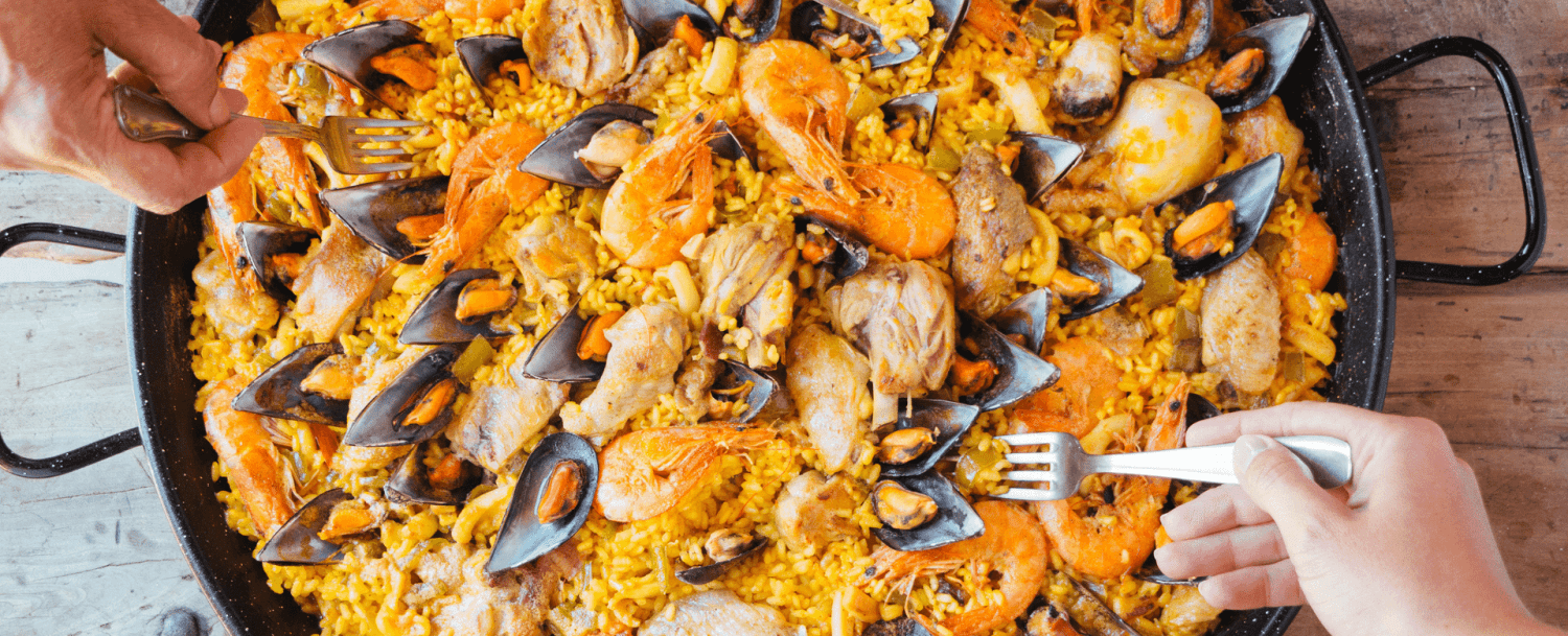 Two hands with forks reaching for a colorful seafood paella.