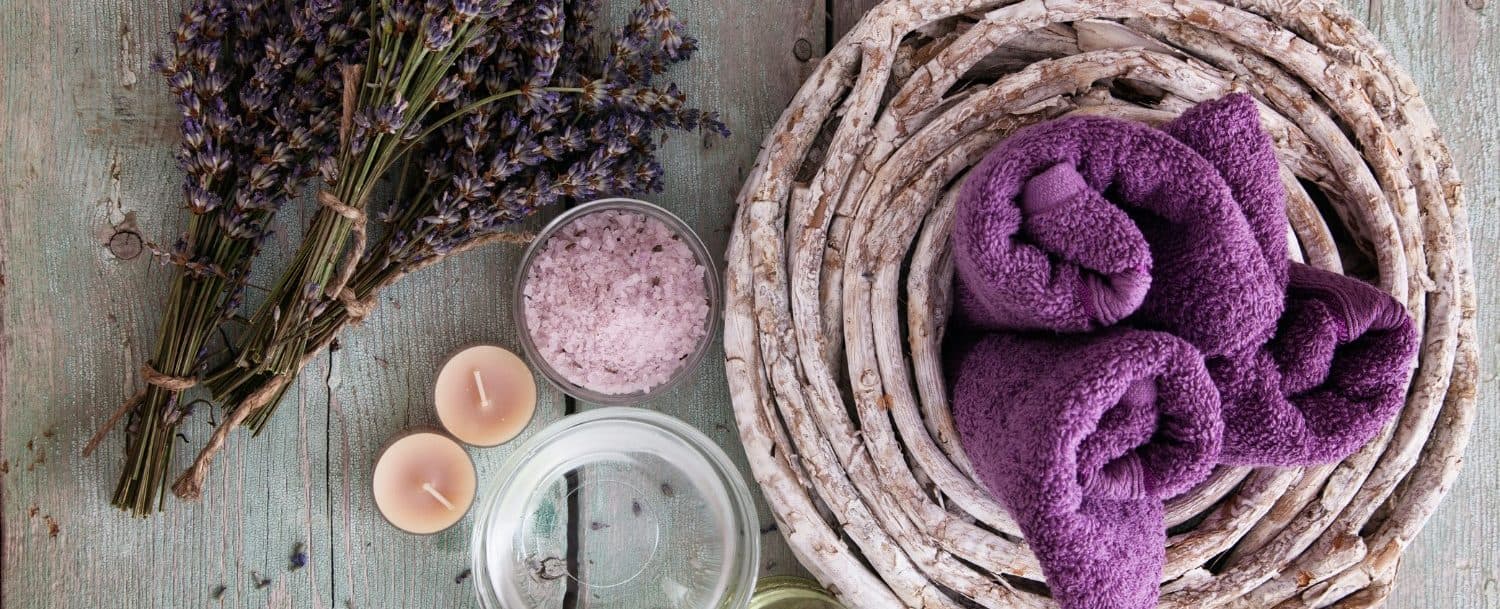 A serene arrangement of lavender, rolled purple towels, salt, candles, and a glass bowl on a rustic wooden surface.