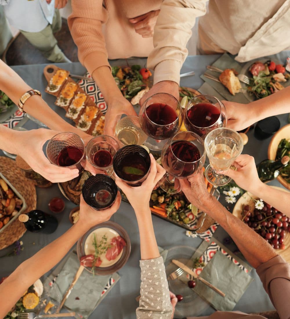 A group of hands raises glasses filled with various drinks in a toast over a festive table.
