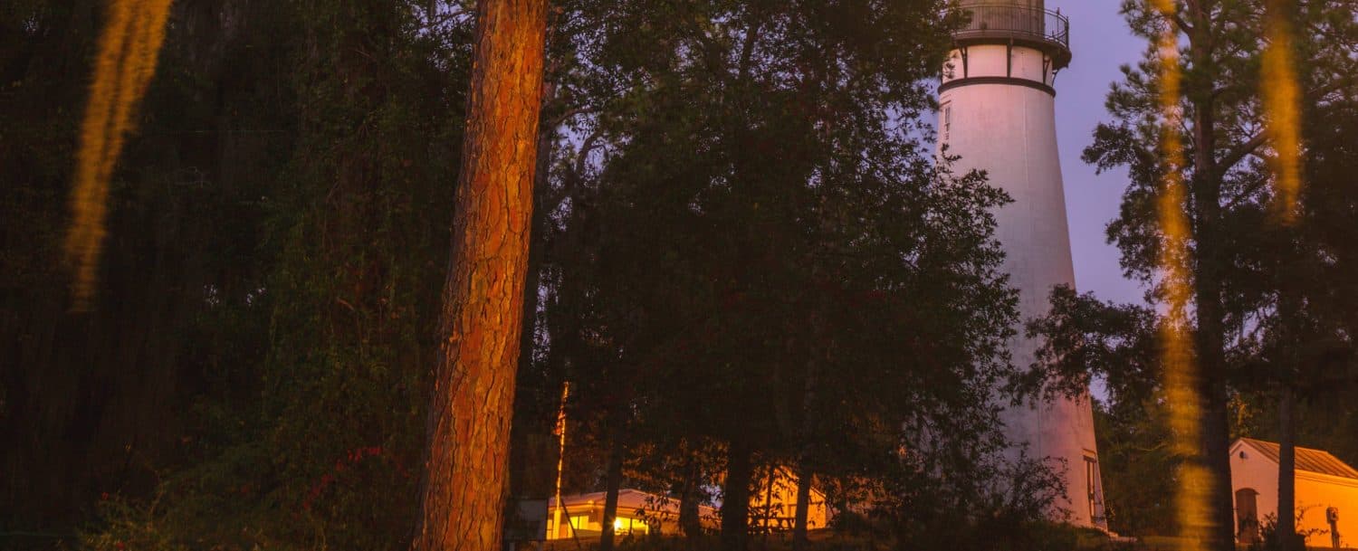 A lighthouse stands among tall trees at dusk, illuminated by warm lights.