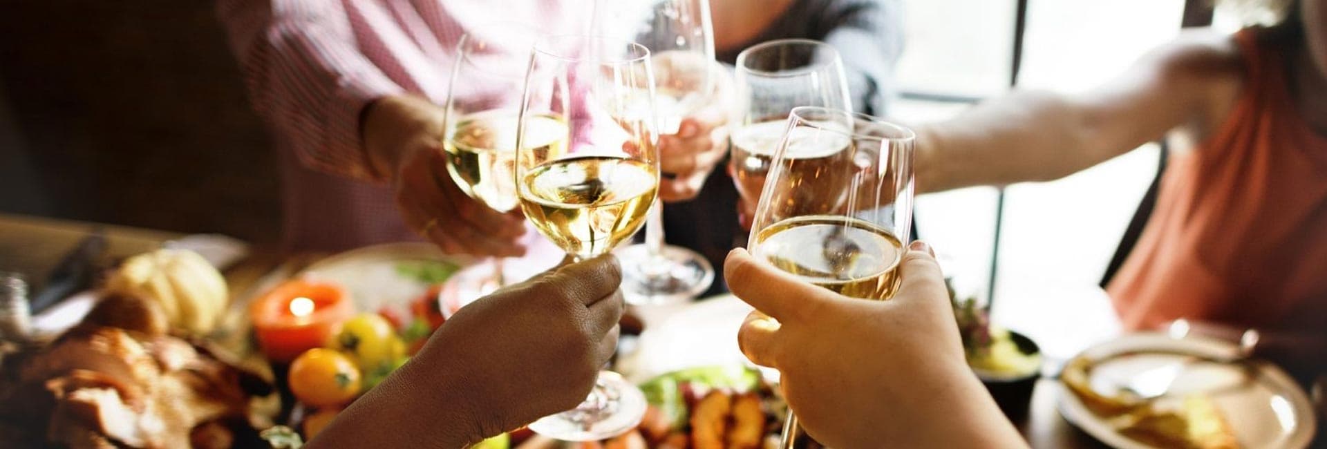 A group of people toasting with glasses of white wine over a table filled with food.