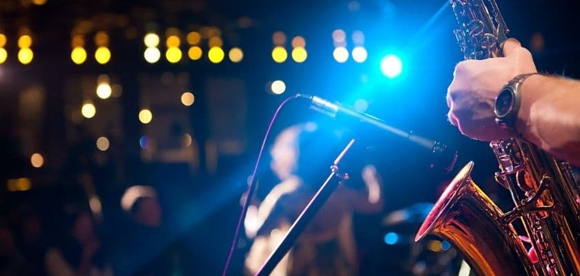 A close-up of a saxophone being played on stage, with colorful lights and an audience in the background.