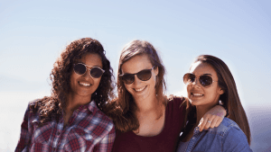 Three women wearing sunglasses smile together against a clear blue sky.