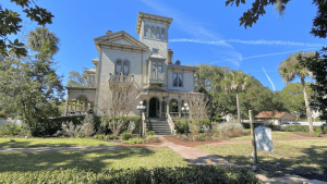 A historic mansion surrounded by greenery and palm trees under a clear blue sky.