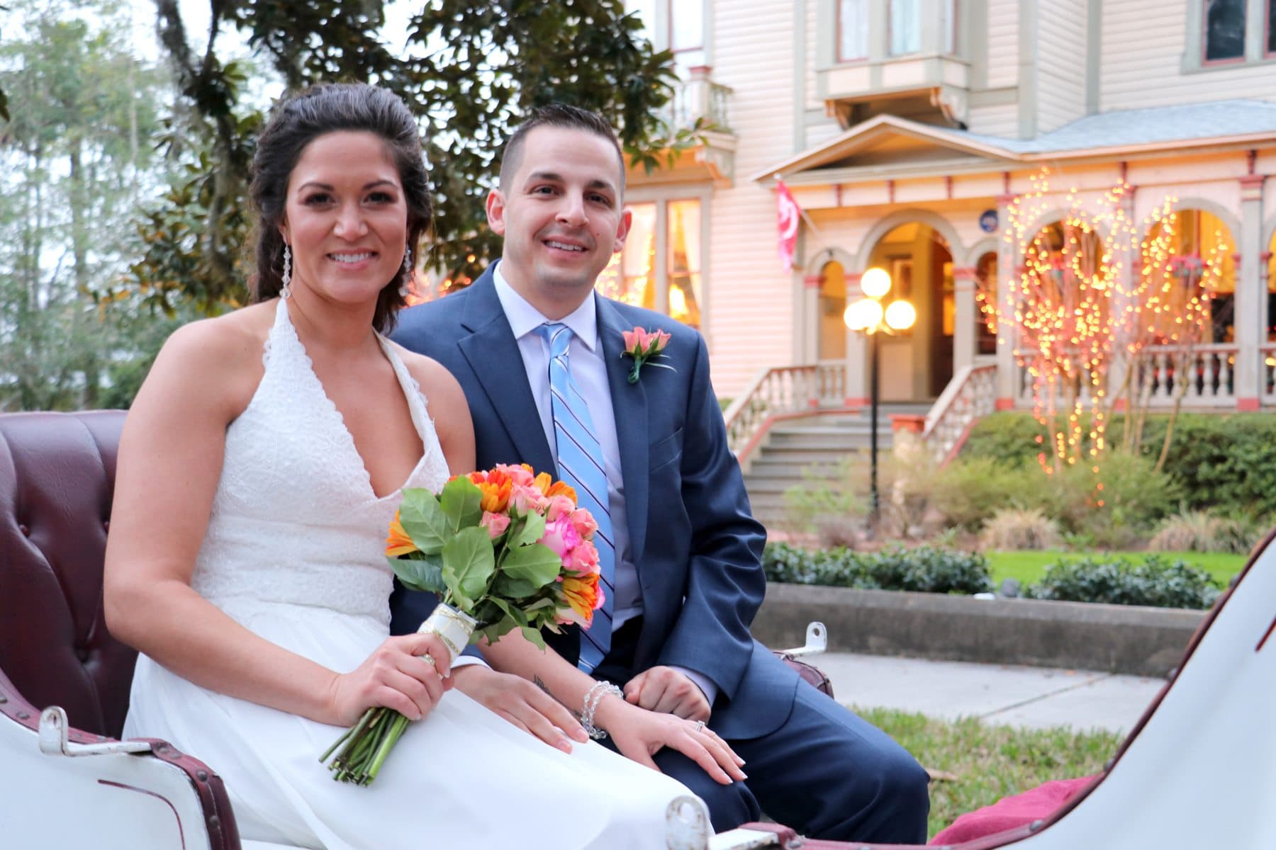 A smiling bride and groom pose together in a carriage, with a beautifully lit historic house in the background.
