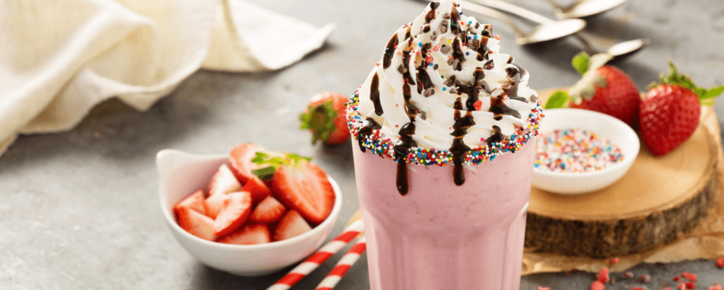 A tall strawberry milkshake topped with whipped cream, chocolate drizzle, and colorful sprinkles, alongside fresh strawberries and a bowl of sprinkles.