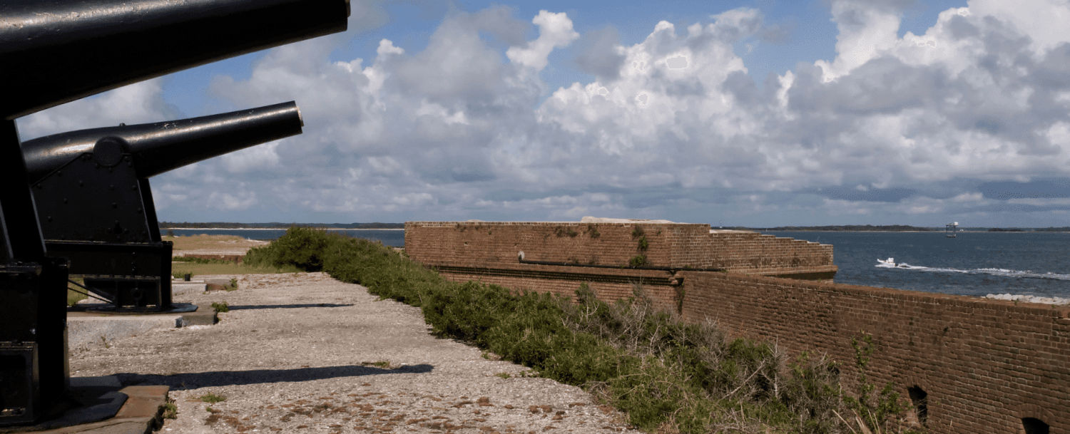 A view of cannons overlooking a coastal landscape with a brick fort and scattered clouds above.