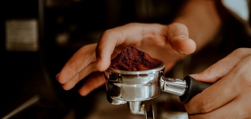 A hand holds a portafilter filled with ground coffee.