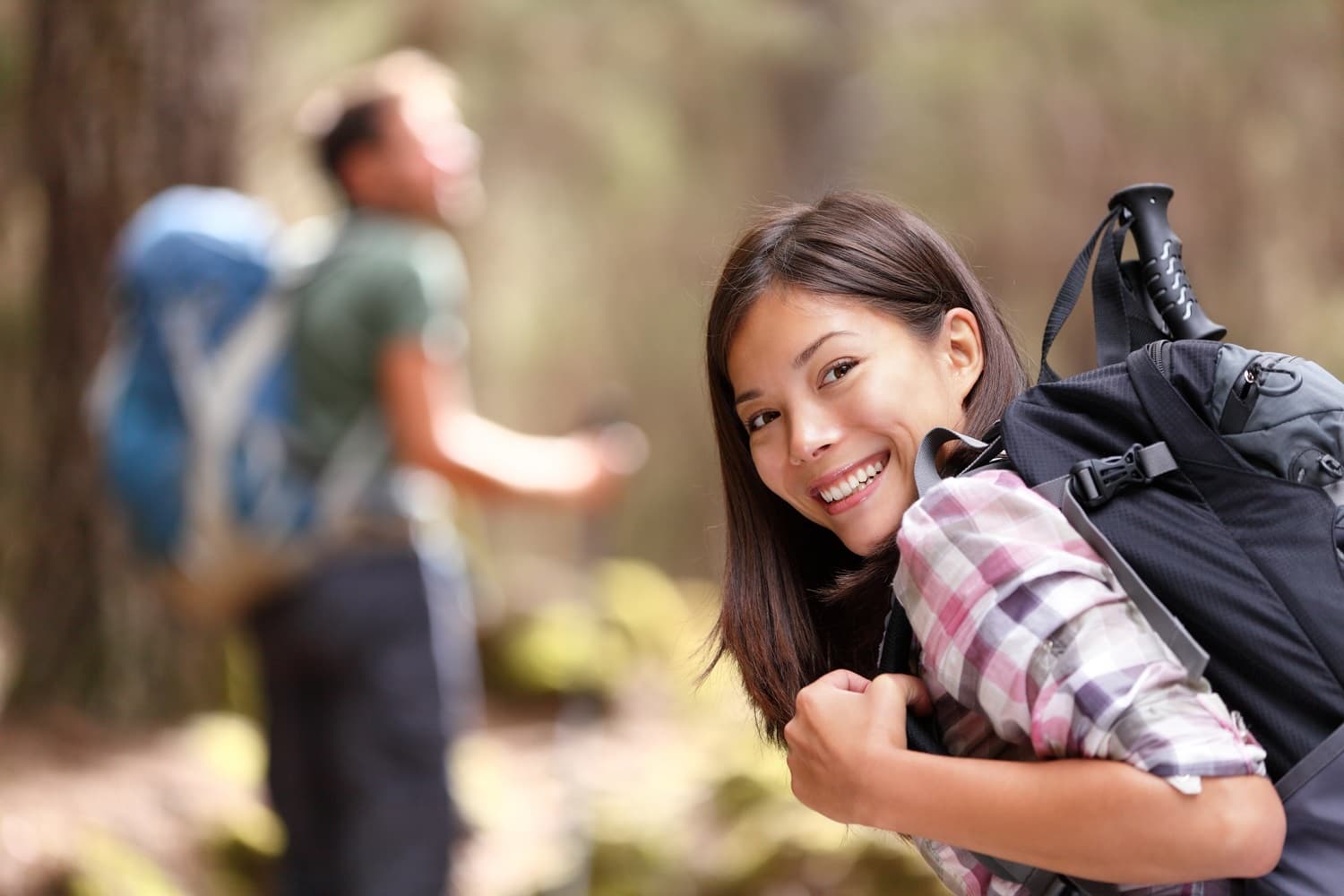 A smiling woman with a backpack poses in the foreground while a man in the background looks at his phone in a forest.