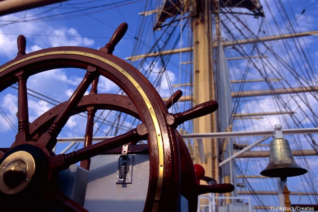 A close-up of a ship's wooden steering wheel against a blue sky.