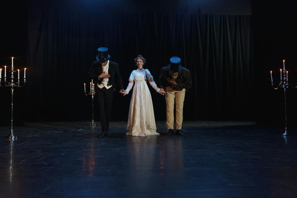 Three performers in formal attire bow on stage with candelabra in the background.