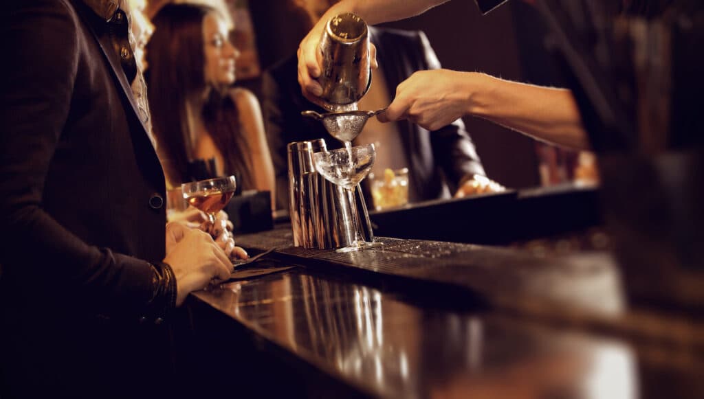 A bartender pours a cocktail into a glass at a bustling bar.