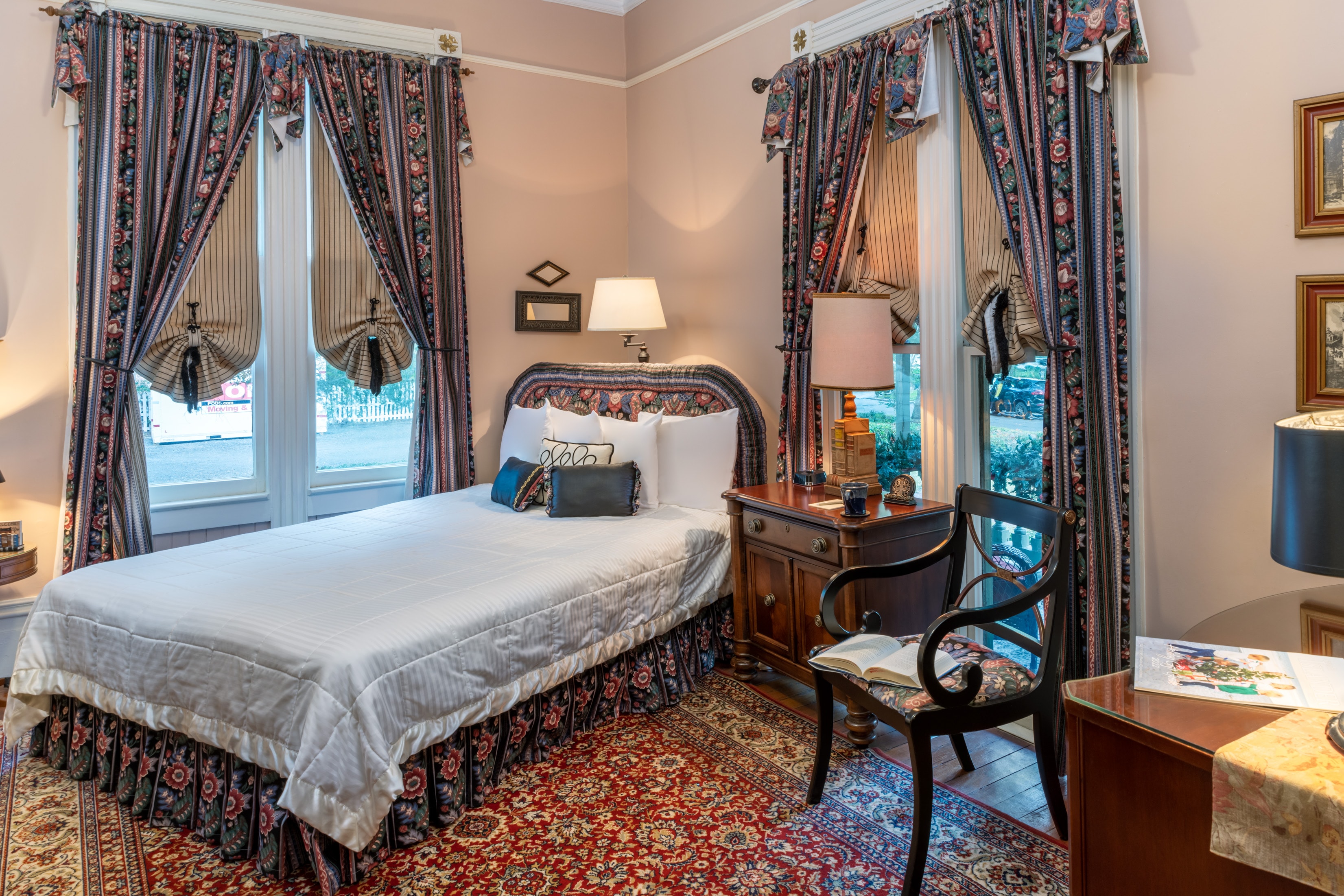 Bright and inviting guest room with a queen bed, antique furnishings, floral drapery, large windows, and classic Southern charm.