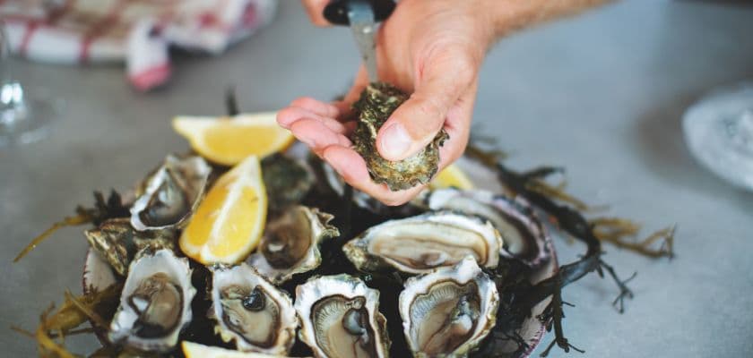 A hand opens an oyster on a platter garnished with lemon wedges.