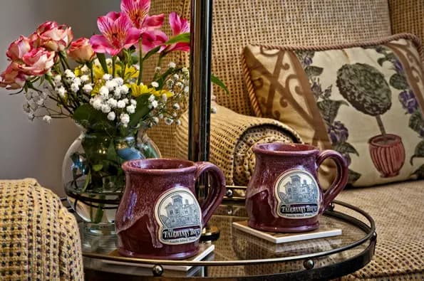 Close-up detail inside a guest suite featuring two signature Fairbanks Inn mugs on a glass-top table beside fresh flowers and a cozy upholstered chair with decorative pillows.