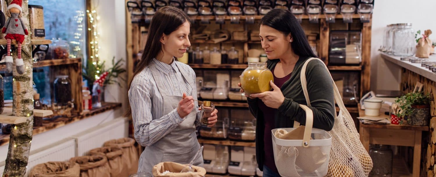 Two women engage in a conversation while shopping in a cozy, eco-friendly store filled with jars and bulk items.