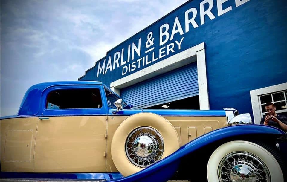 A vintage blue and tan car is parked in front of the Marlin & Barrel Distillery building.