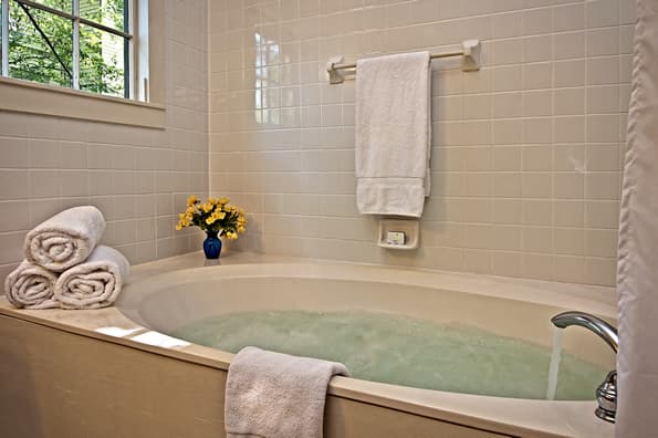 Large, jetted soaking tub with rolled and hanging towels nearby. Flowers in vase on edge of tub