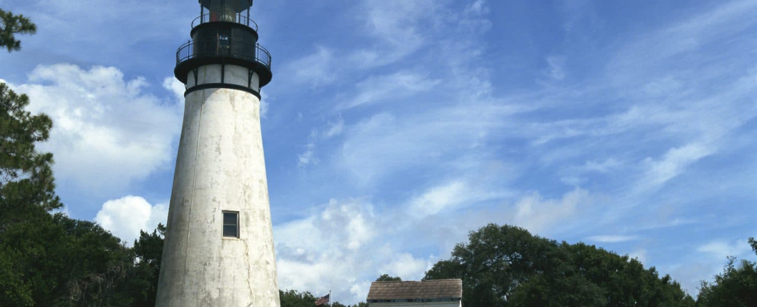 A tall, weathered lighthouse stands beside lush greenery under a blue sky with scattered clouds.