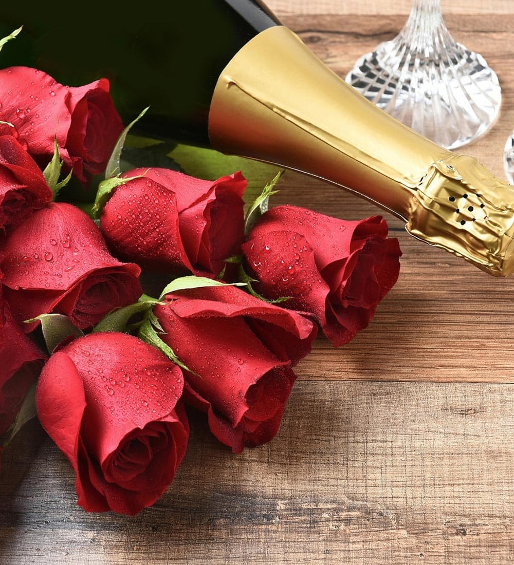 A bottle of champagne beside a cluster of red roses on a wooden surface.