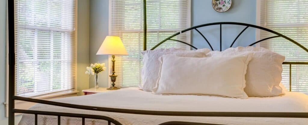 A cozy bedroom featuring a metal-framed bed, soft pillows, a lamp, and natural light from windows.