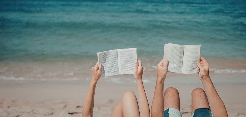 Two people sitting on a beach hold books open, facing the ocean.