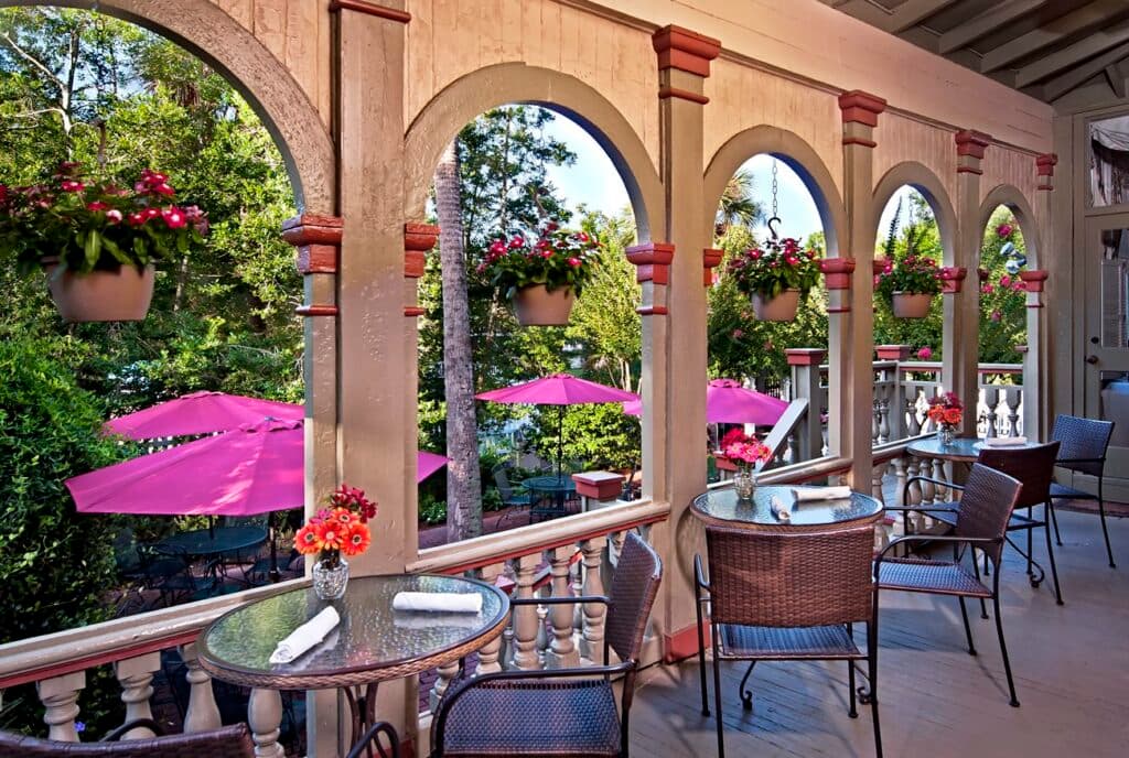 A charming outdoor dining area featuring glass tables, wicker chairs, and vibrant potted flowers under archways, with pink umbrellas in the background.
