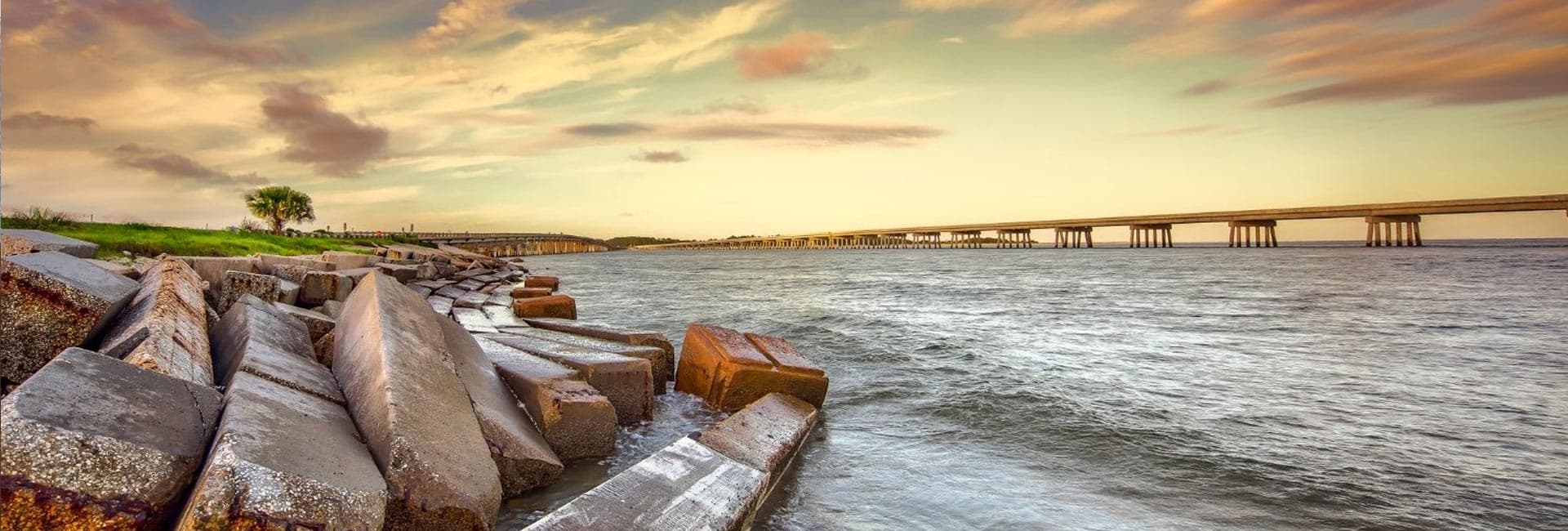 A rocky shoreline with a bridge spanning across the water under a colorful sky at sunset.