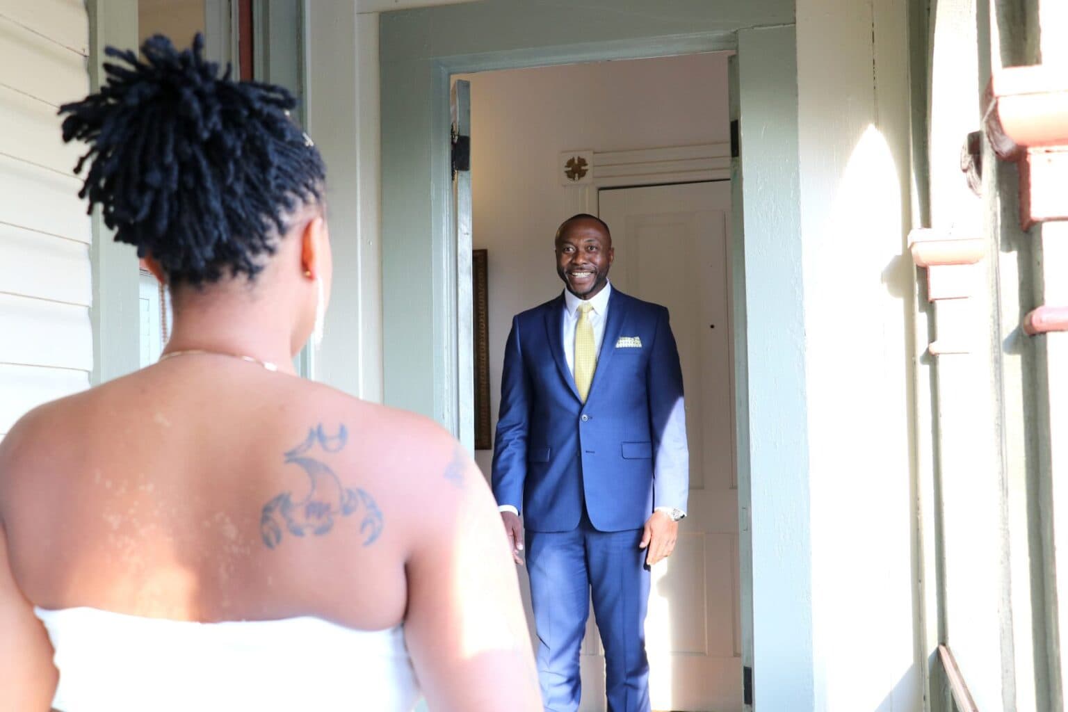 A man in a suit stands at a doorway smiling at a woman with her back turned.
