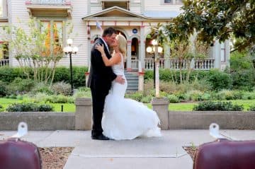 A newlywed couple embraces and kisses in front of a charming house and garden.