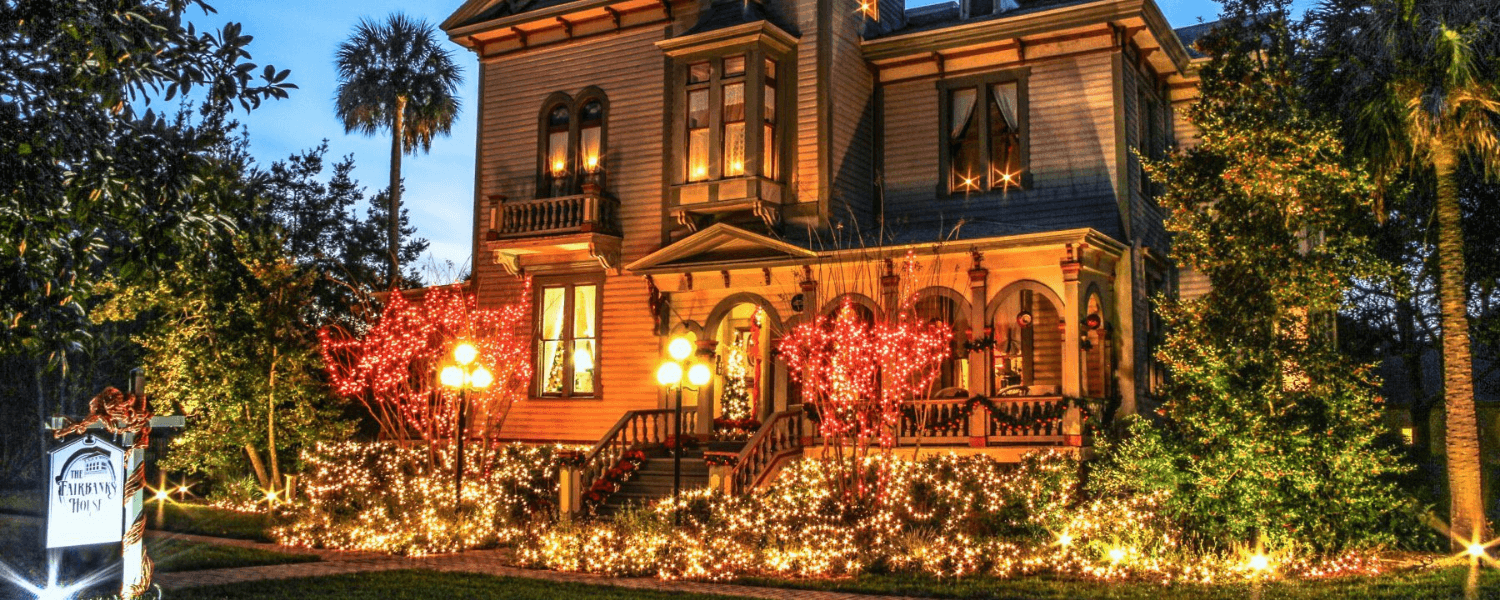 A beautifully lit historic house decorated with festive lights and surrounded by greenery.